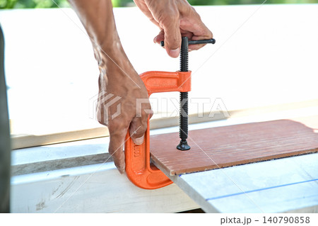 A carpenter's hands tightening a heavy-duty orange metal C-clamp on wood materials during a DIY renovation or construction project. 140790858
