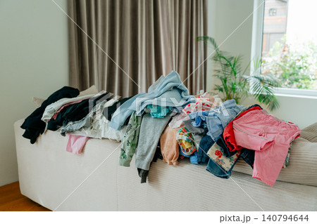 A large, messy pile of colorful baby clothes and garments stacked on a bright sofa in a living room, waiting to be folded or ironed. Stock photo. Housework and laundry concept featuring a big heap of 140794644