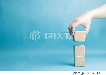 A person's hand carefully placing a blank wooden cube on top of a vertical stack against a minimalist blue backdrop. High Quality Photo A person's hand carefully placing a blank wooden cube on top of a vertical stack against a minimalist blue backdrop. High Quality Photo 140794935