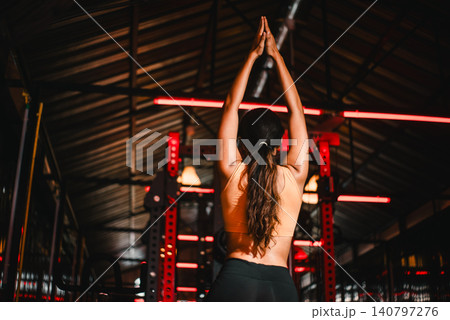 Fitness woman performing standing side stretch with arms overhead in dark gym with red lighting, highlighting flexibility, mobility, warmup, recovery, balance, core strength, and wellness training. Fitness woman performing standing side stretch with arms overhead in dark gym with red lighting, highlighting flexibility, mobility, warmup, recovery, balance, core strength, and wellness training. 140797276