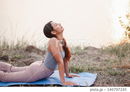 Woman practicing outdoor cobra yoga pose on blue mat near lakeside at sunset, focusing on flexibility, recovery, mindful breathing, back stretch, wellness, relaxation, and healthy lifestyle. Woman practicing outdoor cobra yoga pose on blue mat near lakeside at sunset, focusing on flexibility, recovery, mindful breathing, back stretch, wellness, relaxation, and healthy lifestyle. 140797285