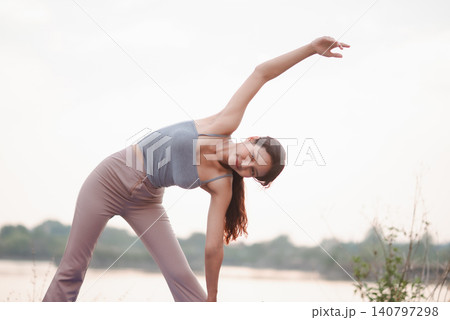 Woman practicing outdoor triangle yoga stretch by lakeside with bright sky, showing flexibility, balance, mobility, mindful breathing, nature wellness, calm focus, and healthy active lifestyle. Woman practicing outdoor triangle yoga stretch by lakeside with bright sky, showing flexibility, balance, mobility, mindful breathing, nature wellness, calm focus, and healthy active lifestyle. 140797298