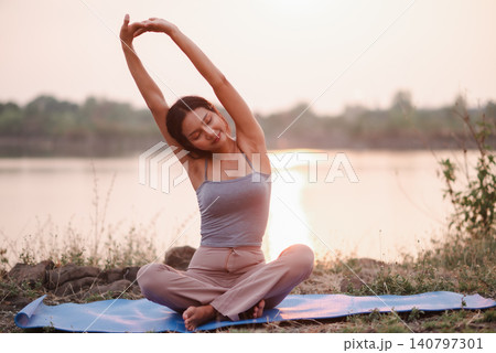 Woman sitting on yoga mat by lakeside sunset, stretching arms overhead with calm smile, showing flexibility, mindful breathing, warmup, relaxation, balance, nature wellness, and peace. Woman sitting on yoga mat by lakeside sunset, stretching arms overhead with calm smile, showing flexibility, mindful breathing, warmup, relaxation, balance, nature wellness, and peace. 140797301