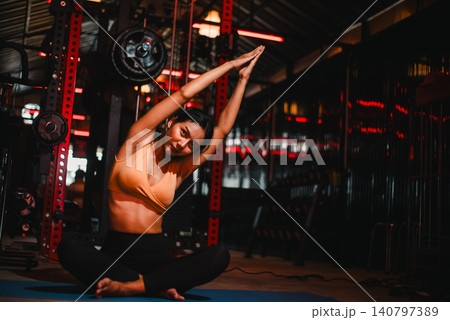 Fitness woman doing seated side stretch on yoga mat in dark gym with red lighting, showing flexibility, mobility, warmup, wellness, workout recovery, balance, and mindful training. 140797389
