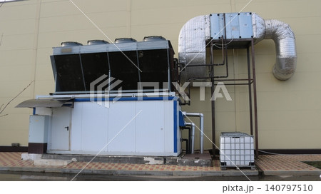 An industrial chiller with a remote condenser is installed against the building wall. The system includes insulated air ducts, cooling fans, and a Eurocube for drainage. The equipment provides climate An industrial chiller with a remote condenser is installed against the building wall. The system includes insulated air ducts, cooling fans, and a Eurocube for drainage. The equipment provides climate 140797510