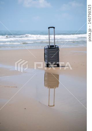 A grey rolling suitcase standing on wet reflective sand on a beach with its mirror image visible below and calm sea and sky in background. Travel and vacation concept A grey rolling suitcase standing on wet reflective sand on a beach with its mirror image visible below and calm sea and sky in background. Travel and vacation concept 140798325