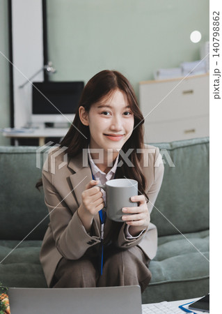 Young asian businesswoman eating a brownie while working from her office on a laptop next to a window with blinds 140798862