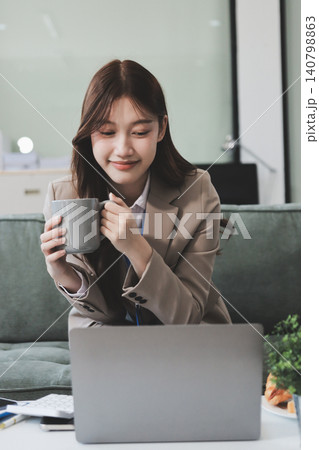 Young asian businesswoman eating a brownie while working from her office on a laptop next to a window with blinds Young asian businesswoman eating a brownie while working from her office on a laptop next to a window with blinds 140798863