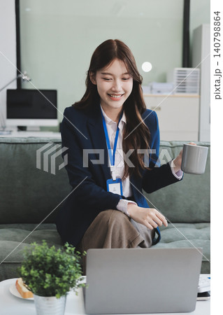 Young asian businesswoman eating a brownie while working from her office on a laptop next to a window with blinds 140798864