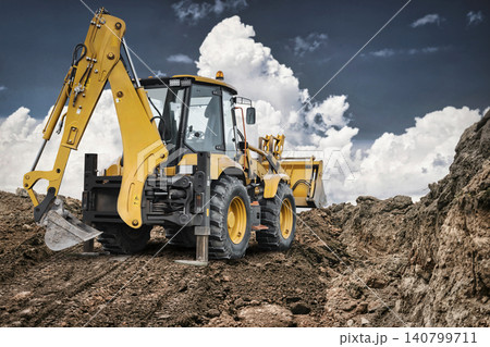 A universal excavator loader operates on a construction site, moving dirt. The sky is cloudy. Heavy machinery is at work in the background 140799711