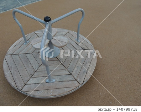 The children's round carousel is installed on a safe rubber surface. The structure consists of a wooden plank deck, a central metal support, comfortable handrails, and three petal-shaped seats. 140799718