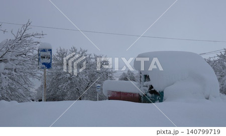 A snow-covered truck sits at a bus stop after a heavy snowfall. Huge drifts cover the truck's cabin, a road sign, and tree branches. The atmosphere is reminiscent of a frosty winter day. A snow-covered truck sits at a bus stop after a heavy snowfall. Huge drifts cover the truck's cabin, a road sign, and tree branches. The atmosphere is reminiscent of a frosty winter day. 140799719