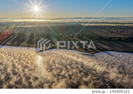 Aerial view of fog drifting across Lake Keepit Dam at sunrise as golden light casts long shadows across the surrounding rural landscape and reservoir in NSW, Australia. 140800181