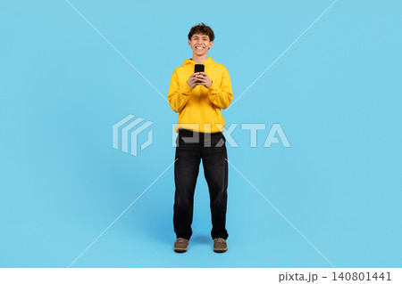 Smiling young guy in yellow hoodie standing with smartphone on blue background 140801441