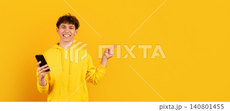 Smiling guy in yellow hoodie holding smartphone and pointing to empty space Smiling guy in yellow hoodie holding smartphone and pointing to empty space 140801455