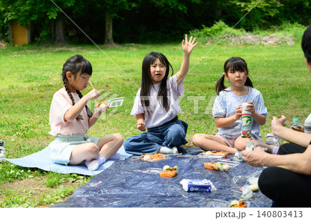 新緑の公園の広場でレジャーシートを広げ食事の前に挨拶をする女子小学生たち 新緑の公園の広場でレジャーシートを広げ食事の前に挨拶をする女子小学生たち 140803413