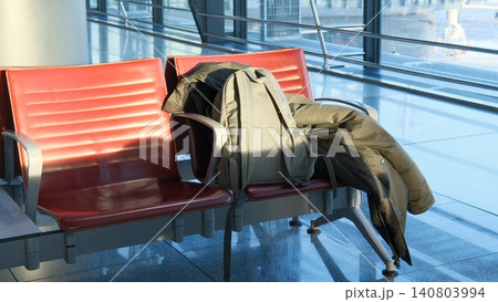 Waiting area at airport, empty chairs with backpack and jacket on bench in hall. Flight delay, luggage, carry-on baggage before departure. Duty free zone. Waiting for boarding at gate Waiting area at airport, empty chairs with backpack and jacket on bench in hall. Flight delay, luggage, carry-on baggage before departure. Duty free zone. Waiting for boarding at gate 140803994
