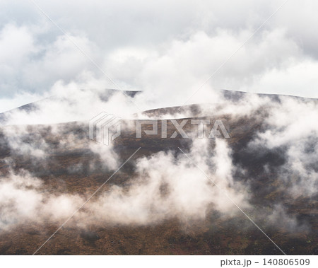 Mist drifts across a brown, heather-covered mountainside under a bright sky. Wisps of white fog obscure the tops of distant, rugged hills. 140806509
