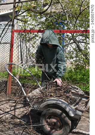 Teen boy carrying branches toward metal garden trailer in rural yard near fence. 140807470