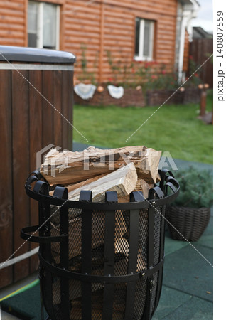 Wooden outdoor hot tub with firewood basket standing on terrace near country house. Hot tub used for relaxation in backyard. Traditional wooden hot tub heated by fire in garden. vertical Wooden outdoor hot tub with firewood basket standing on terrace near country house. Hot tub used for relaxation in backyard. Traditional wooden hot tub heated by fire in garden. vertical 140807559