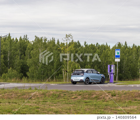 Modern blue compact electric vehicle charging at a public EV station in a green park environment next to a forest. Modern blue compact electric vehicle charging at a public EV station in a green park environment next to a forest. 140809564