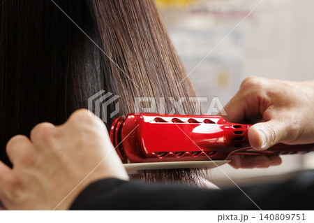 赤いヘアアイロンで女性の髪をストレートにする美容師 140809751
