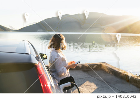 Woman traveler with charging plug next to her electric car on the sea coast at sunset Woman traveler with charging plug next to her electric car on the sea coast at sunset 140810042