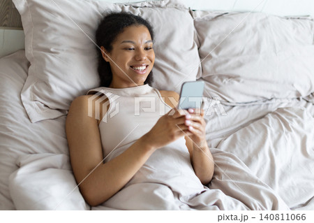 Happy black woman lying in bed and using smartphone, smiling while texting Happy black woman lying in bed and using smartphone, smiling while texting 140811066