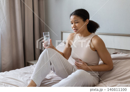 Black woman is sitting on bed holding glass of water and touching her stomach Black woman is sitting on bed holding glass of water and touching her stomach 140811100