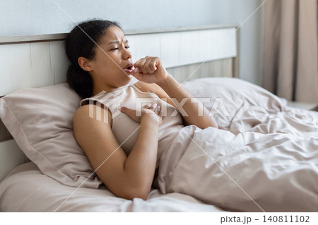 Black woman lying in bed and coughing with one hand on chest 140811102