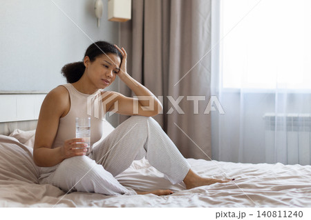 Black woman sitting on bed, holding glass of water and touching head 140811240