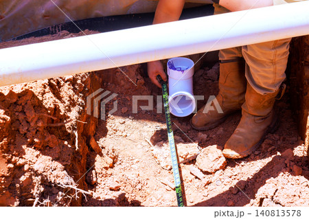Contractor works at construction site, measuring pipe in trench with tools equipment present. Contractor works at construction site, measuring pipe in trench with tools equipment present. 140813578