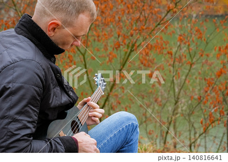 Man plays ukulele while spending time outdoors in autumnal natural setting. Musician holds small string instrument and performs music in relaxed atmosphere surrounded by greenery and open space Man plays ukulele while spending time outdoors in autumnal natural setting. Musician holds small string instrument and performs music in relaxed atmosphere surrounded by greenery and open space 140816641
