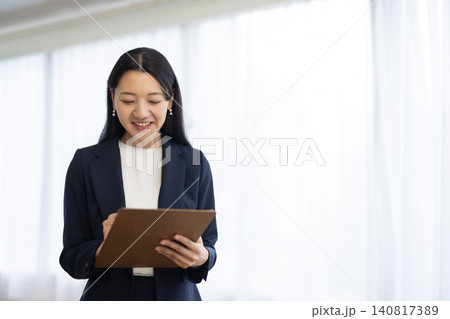 窓辺でバインダーにメモをとりながら微笑む日本人の若いビジネスウーマン 窓辺でバインダーにメモをとりながら微笑む日本人の若いビジネスウーマン 140817389