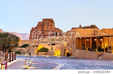 View of the historic mud brick architecture and sandstone mountains in AlUla Old Town, Saudi Arabia, featuring a modern walkway with ambient lighting at dusk. View of the historic mud brick architecture and sandstone mountains in AlUla Old Town, Saudi Arabia, featuring a modern walkway with ambient lighting at dusk. 140818344