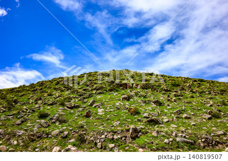 Lush green slope with rocks under clear blue sky with clouds 140819507