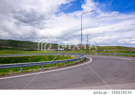 Winding road with guardrail and streetlight in grassy hills under blue sky Winding road with guardrail and streetlight in grassy hills under blue sky 140819534
