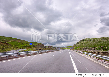 Straight road winds through grassy hills under heavy cloud cover Straight road winds through grassy hills under heavy cloud cover 140819540