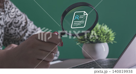 HR and Recruitment concept: Hand holding a magnifying glass over a digital resume profile icon near a laptop 140819938
