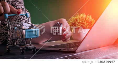 Online real estate shopping concept. Woman holding miniature house near laptop and shopping cart. Online real estate shopping concept. Woman holding miniature house near laptop and shopping cart. 140819947