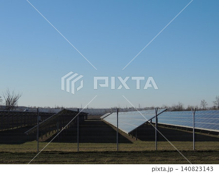 An industrial solar power plant with rows of ground-mounted silicon photovoltaic modules. The metal structures are oriented at a fixed angle to the sun. The site is enclosed by a utility perimeter. 140823143
