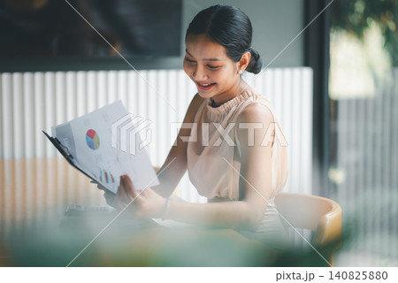 Smiling Asian businesswoman reviewing financial charts in modern cafe office, holding report with confidence, representing accounting success, budget analysis, corporate growth, and productivity. 140825880