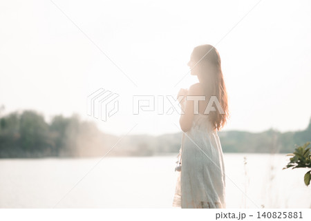 Soft backlit Asian woman in white lace dress standing beside a lake with clasped hands, showing prayer, gratitude, hope, mindfulness, peaceful nature, spiritual calm, and wellness. Soft backlit Asian woman in white lace dress standing beside a lake with clasped hands, showing prayer, gratitude, hope, mindfulness, peaceful nature, spiritual calm, and wellness. 140825881