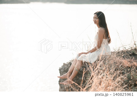 Asian woman in white lace dress sitting on rocky lakeshore in soft sunlight, enjoying quiet nature, calm reflection, peaceful solitude, relaxation, wellness, beauty, and outdoor lifestyle. Asian woman in white lace dress sitting on rocky lakeshore in soft sunlight, enjoying quiet nature, calm reflection, peaceful solitude, relaxation, wellness, beauty, and outdoor lifestyle. 140825961