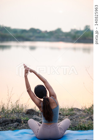 Back view Asian woman practicing seated yoga side stretch on blue mat by a calm lake at sunset, peaceful outdoor wellness, mindful breathing, flexibility training, nature relaxation and self care. Back view Asian woman practicing seated yoga side stretch on blue mat by a calm lake at sunset, peaceful outdoor wellness, mindful breathing, flexibility training, nature relaxation and self care. 140825963