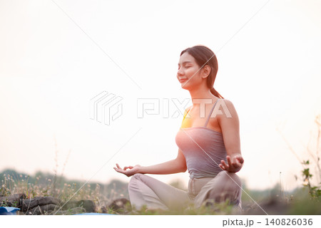 Asian woman meditating on blue yoga mat outdoors by a soft sunset lake, seated lotus pose with mudra hands, peaceful mindfulness, calm breathing, wellness, relaxation and self care. Asian woman meditating on blue yoga mat outdoors by a soft sunset lake, seated lotus pose with mudra hands, peaceful mindfulness, calm breathing, wellness, relaxation and self care. 140826036