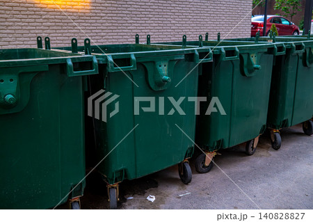 Row of large green industrial waste containers parked along brick wall on city street, urban waste management concept Row of large green industrial waste containers parked along brick wall on city street, urban waste management concept 140828827
