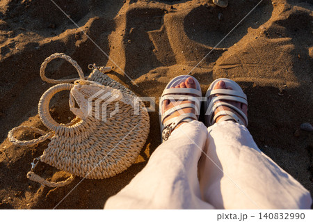 POV shot of woven straw bag and feet in sandals on beach sand 140832990