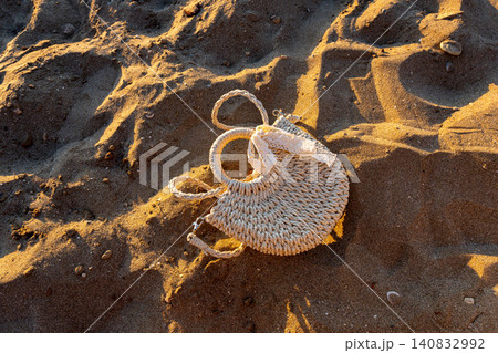 Woven straw bag with lace ribbon on golden beach sand at sunset Woven straw bag with lace ribbon on golden beach sand at sunset 140832992