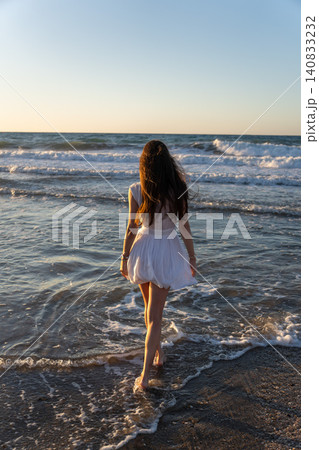 Full length rear view of young woman walking barefoot in sea foam on Crete beach Full length rear view of young woman walking barefoot in sea foam on Crete beach 140833232